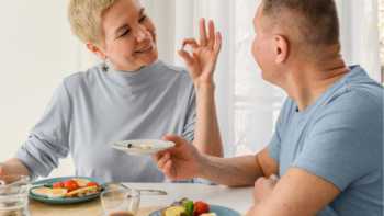 Woman holding a pill and showing it to a man while sitting at a table about to eat a meal