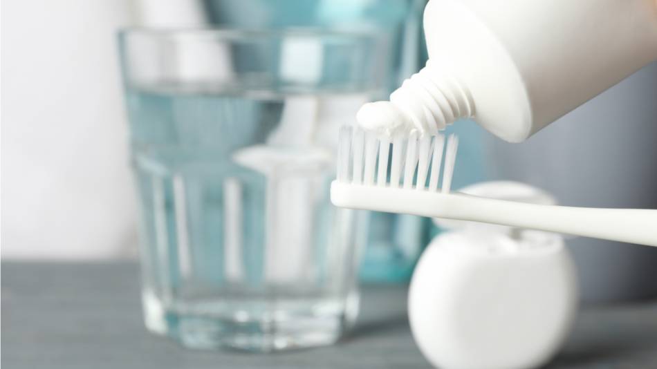 Small amount of toothpaste being applied to a toothbrush with a glass of water in the background