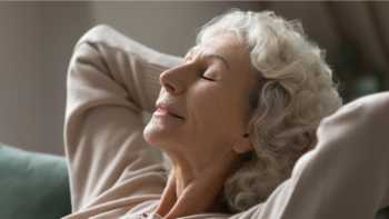 Older women sitting calmly on a couch with her hands behind her head and her eyes closed