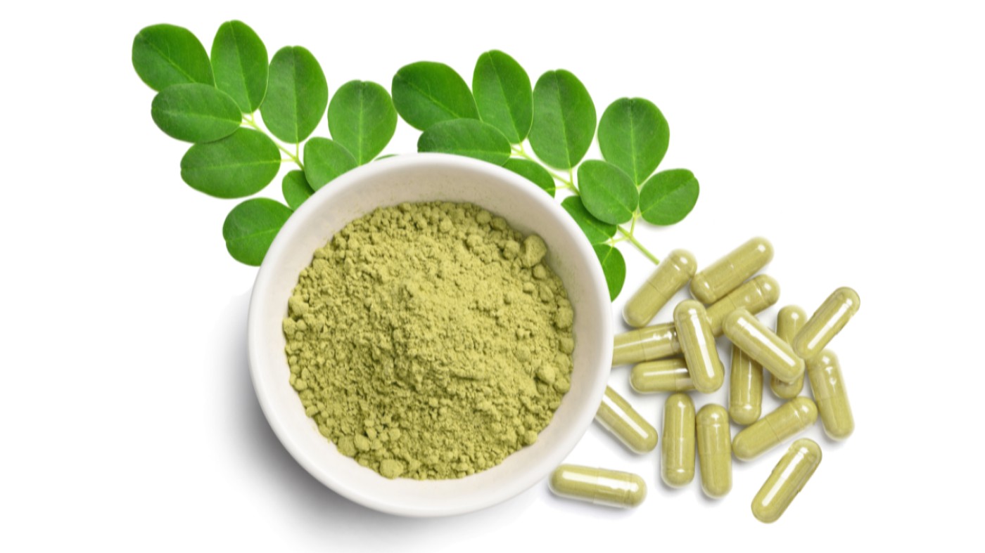 Fresh moringa leaves under a white bowl containing moringa leaf powder that is next to capsules of moringa leaf